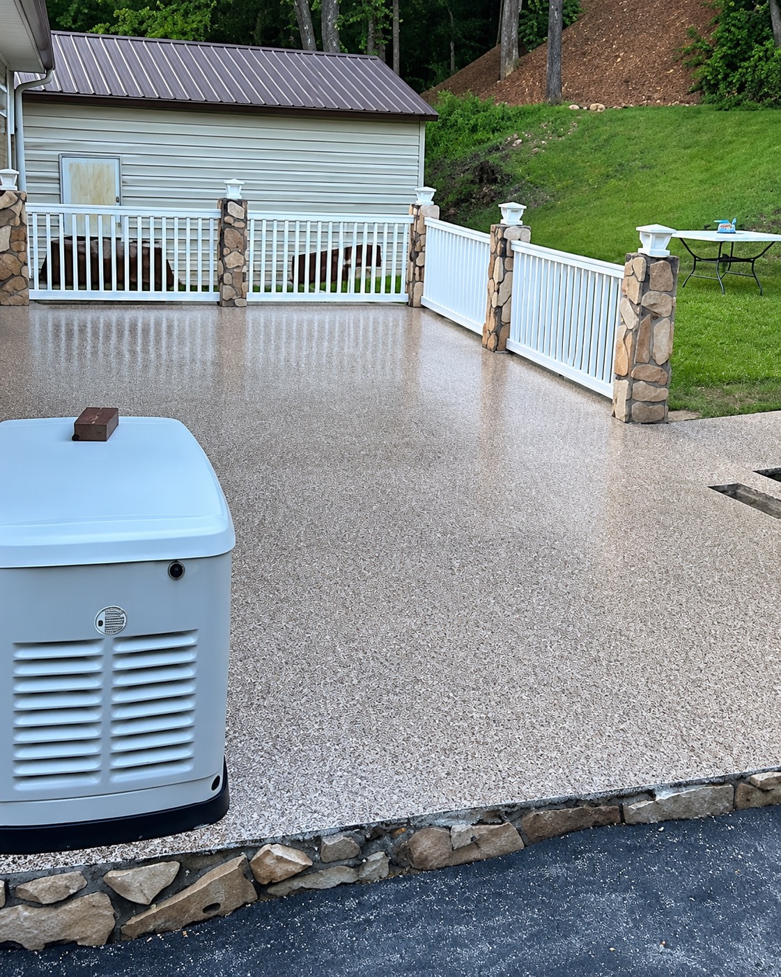 Outdoor patio with a tan flake coating, white vinyl railing between stone-veneer pillars, above a stone border