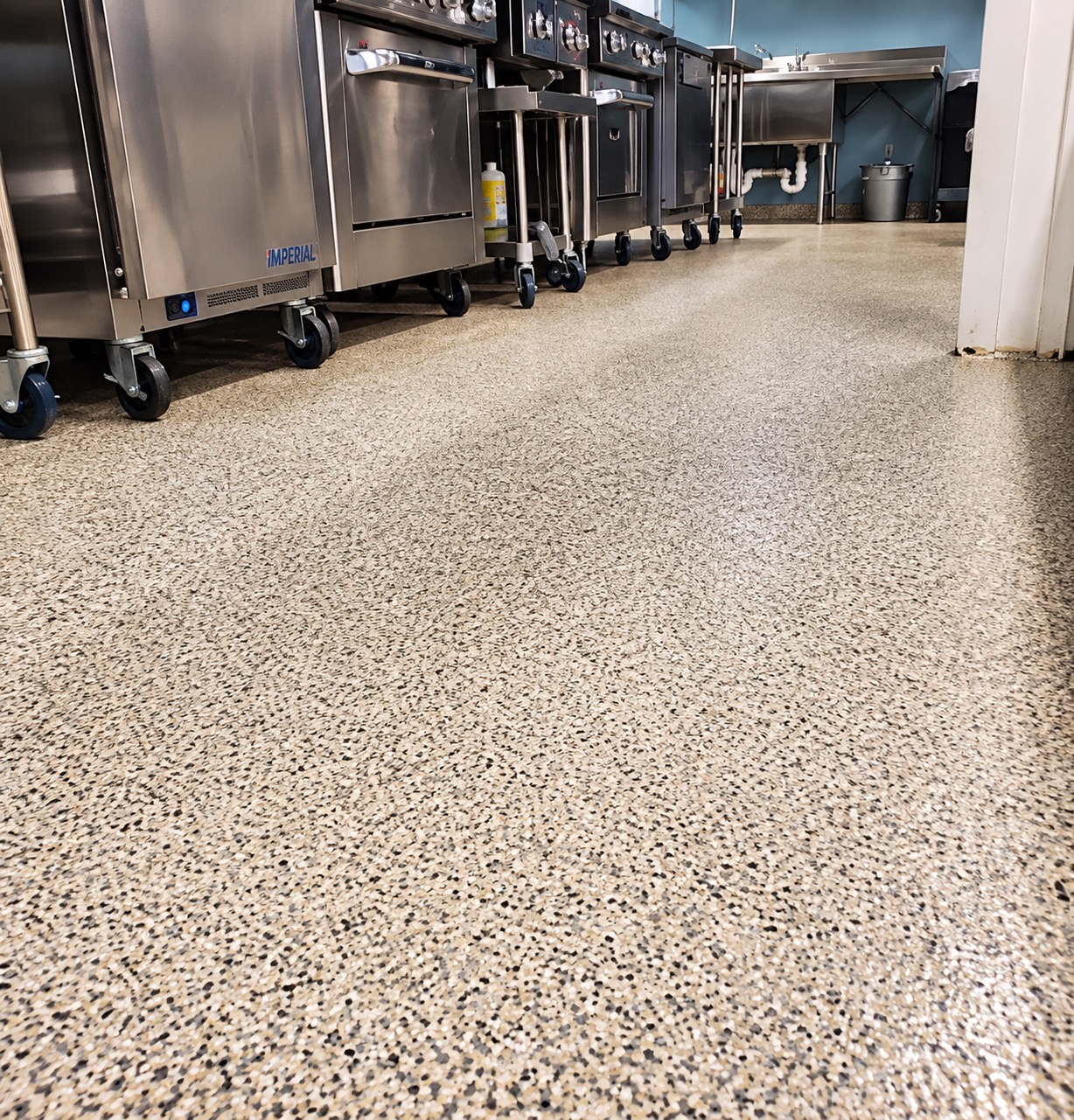 Commercial kitchen with a tan flake floor beneath stainless steel ovens and prep equipment on casters
