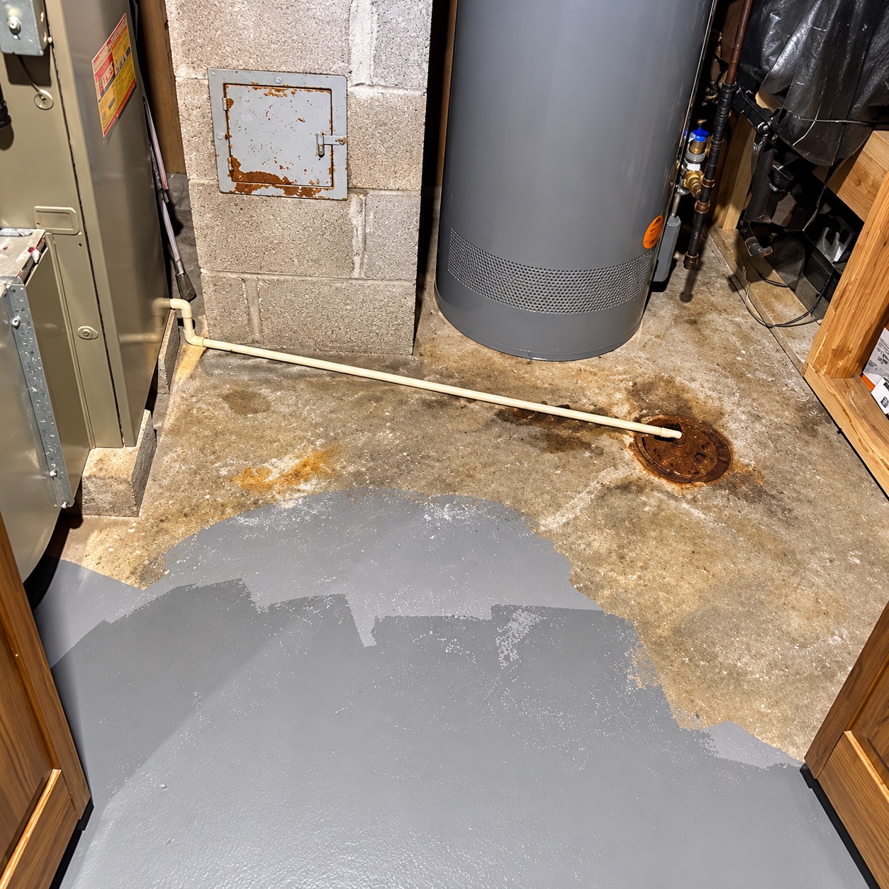 Basement utility room mid-job: rust and water stains around the floor drain, with a gray base coat partly applied near the doorway