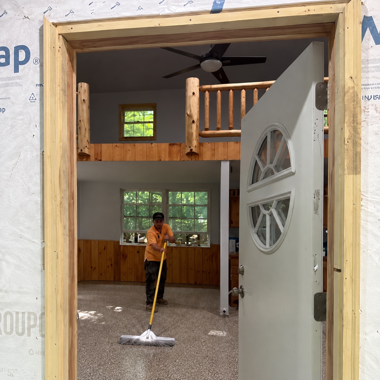Crew member sweeping the flake floor in a cabin great room with a log loft railing, seen through the open front door