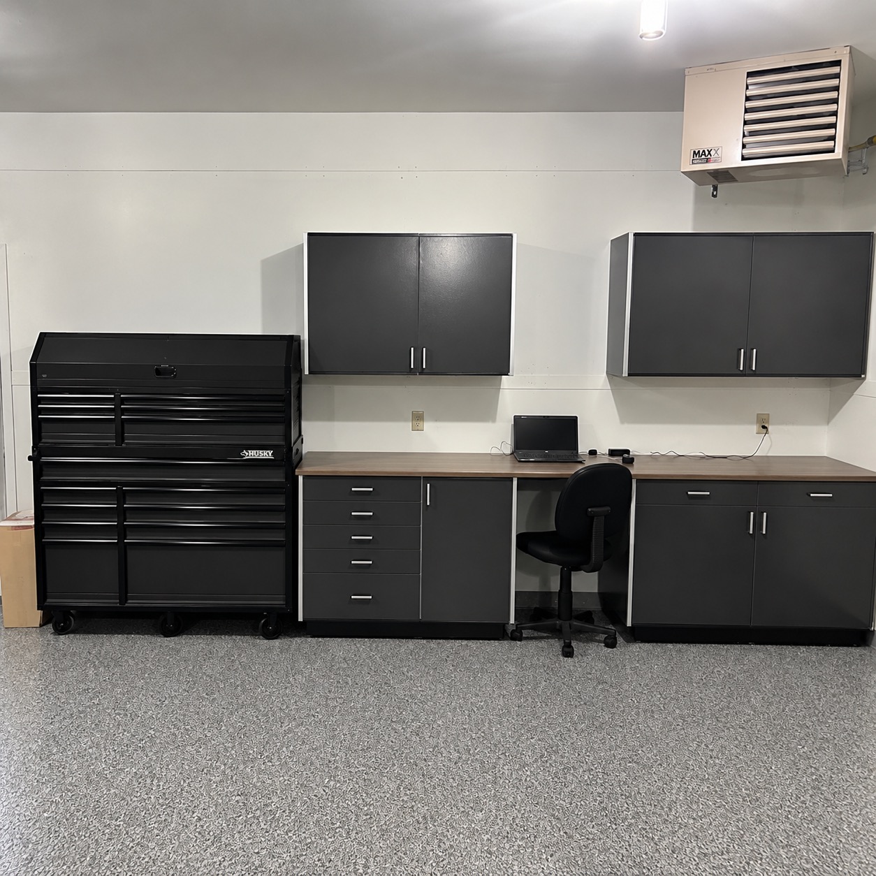 Garage with a gray flake-coated floor beneath gray storage cabinets, a workbench desk and a black rolling tool chest